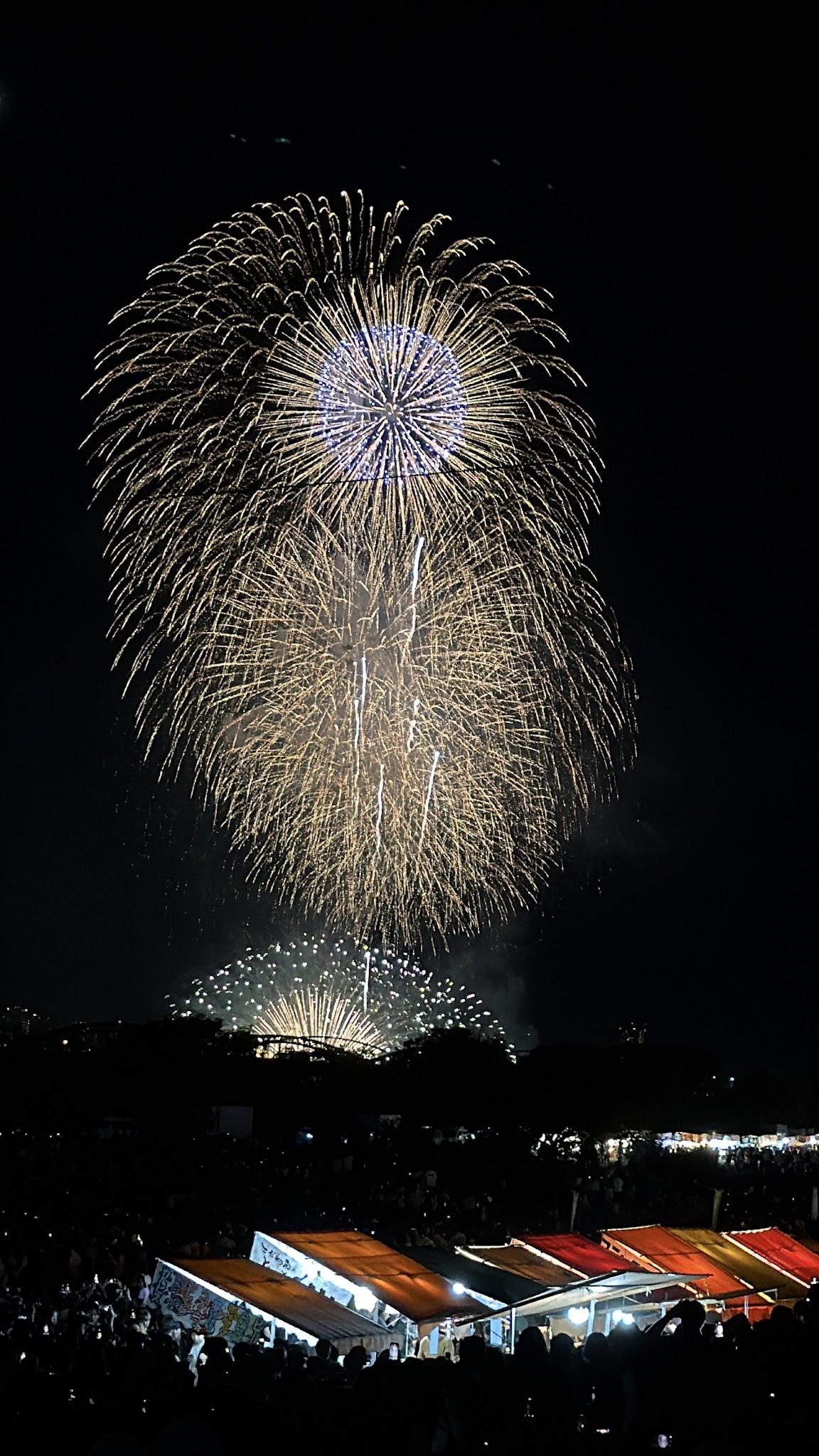 藍心金色煙火在夜空中上下交疊，下方是河岸人群與攤位