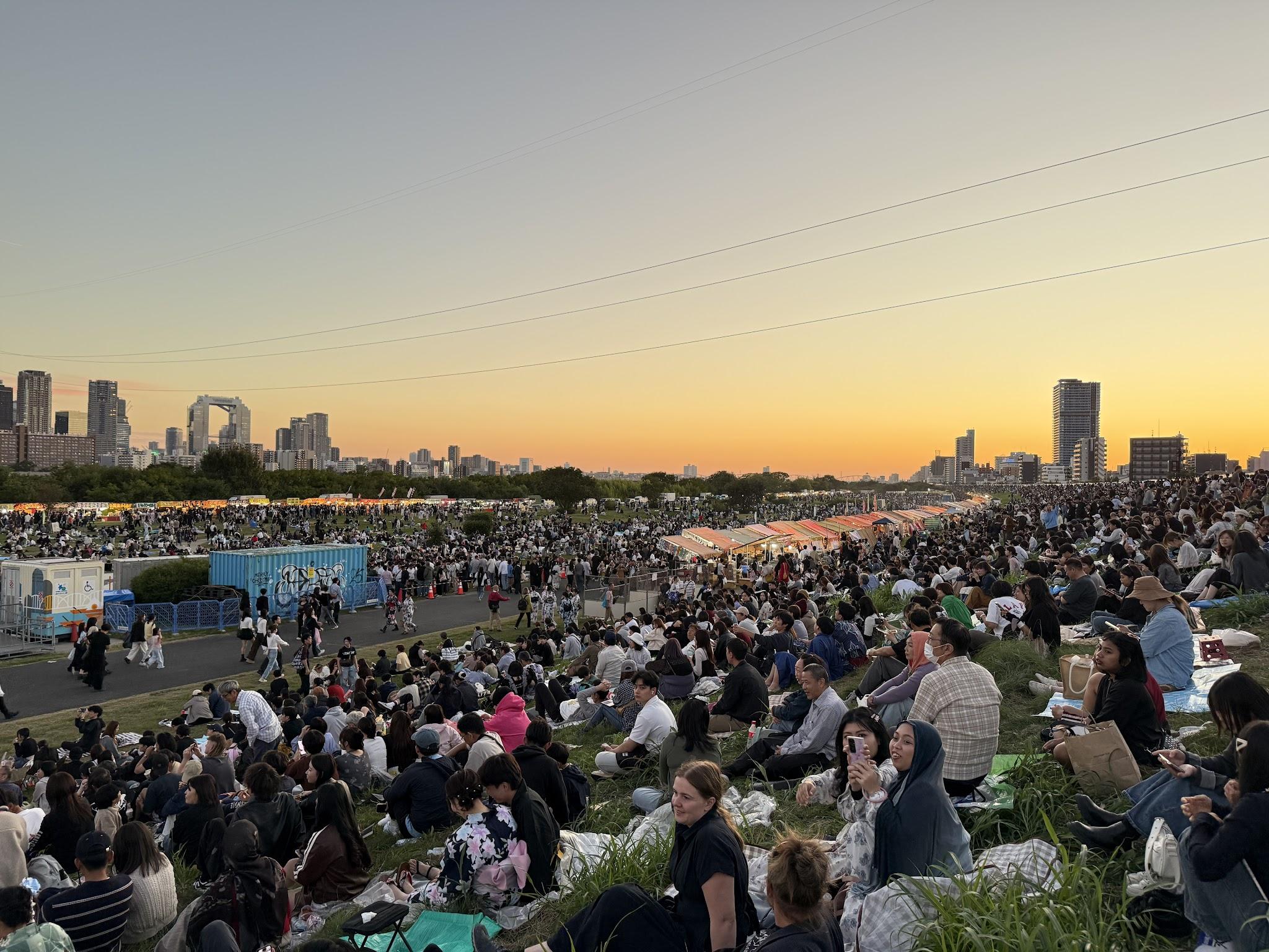 淀川河畔广闊草地上密集坐著等待煙火的人群，遠方是大阪城市天際線